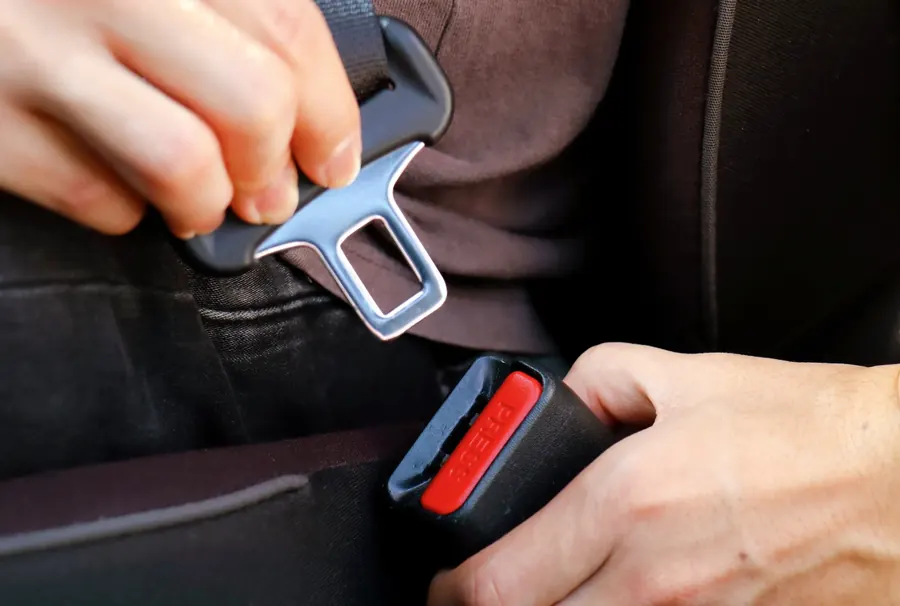 Two hands guiding a metal seatbelt latch plate towards a black buckle receiver with a red release button.