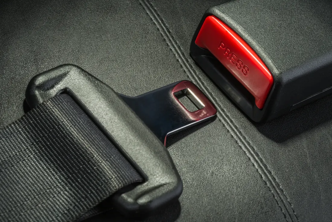 A seatbelt latch plate on black webbing resting beside its red-release buckle receiver on a dark leather vehicle seat.