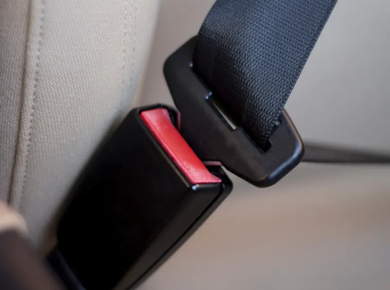 A black seatbelt buckle receiver with a red release button, positioned beside a latch plate on a cream fabric seat.
